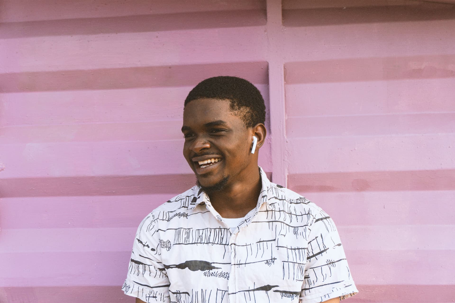 Person in a patterned shirt against a pink wall wearing a wireless earpiece.