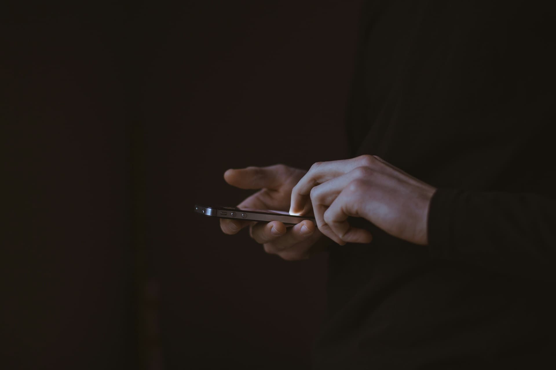 Person in dark clothing using a smartphone against a dim background.