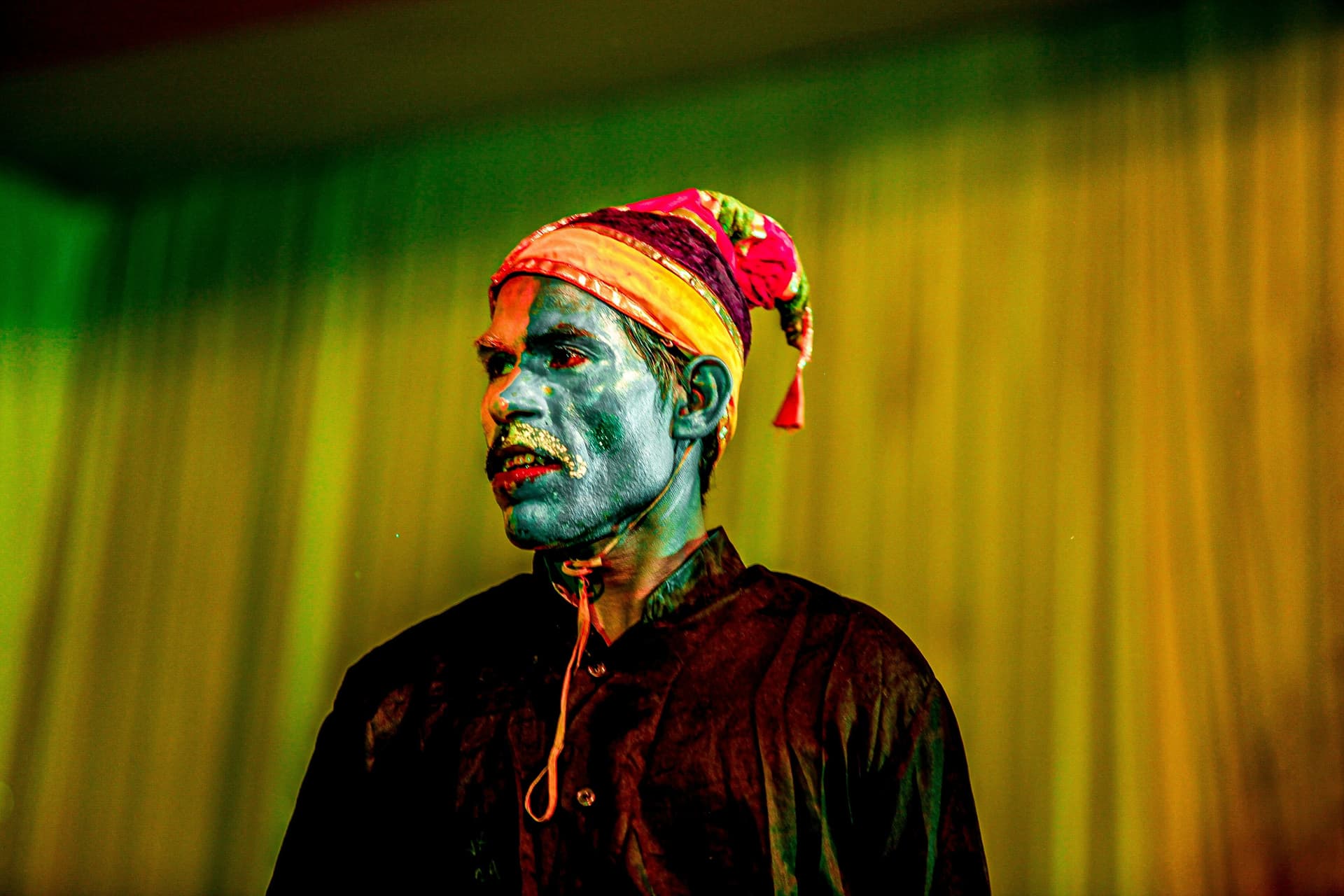 A man with blue face paint and a colorful traditional hat stands against a softly lit background. He wears a black shirt and displays a serious expression, embodying a theatrical performance.